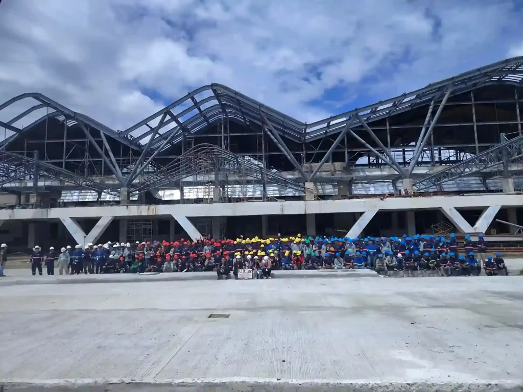 A large group of construction workers in safety gear gathered in front of a steel-framed building under construction