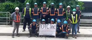 A group of construction workers in PPE posing outdoors with a company banner near greenery and a metal railing.