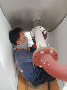Worker in confined space inspecting a pipeline flange with safety glasses and face covering.