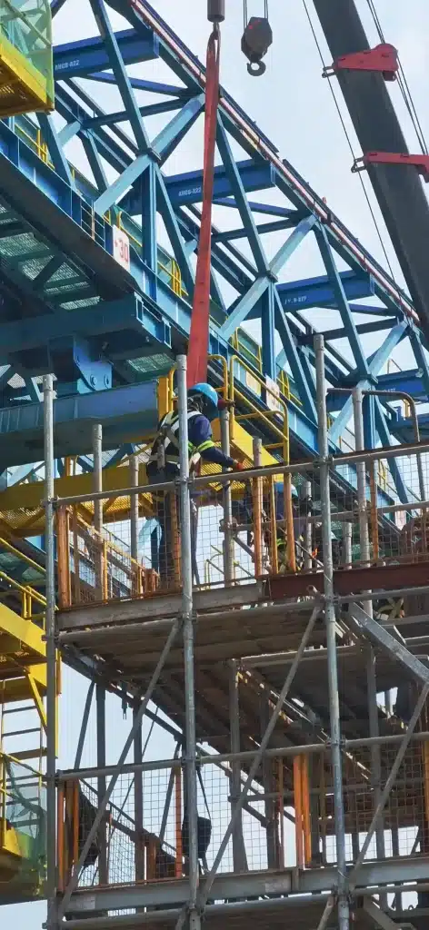 A construction worker on scaffolding handling a crane strap near a steel framework with safety gear.