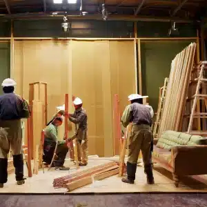 Five workers in white helmets handling wooden planks in an indoor carpentry space with ladders and a couch